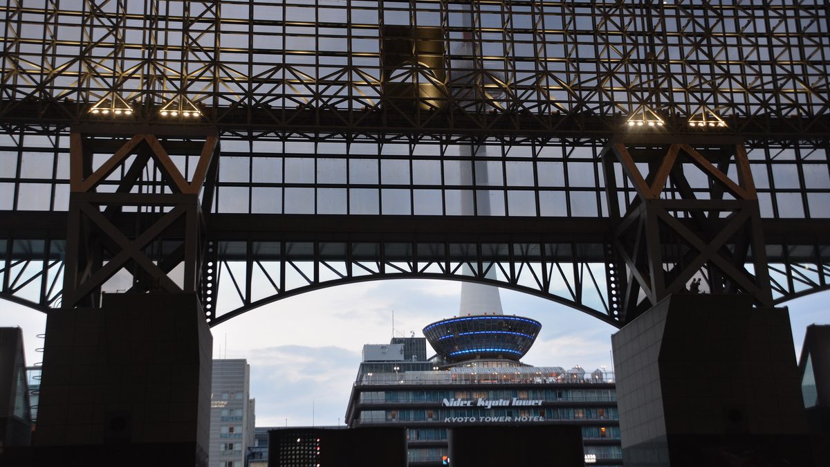 Kyoto Tower lit at dusk, framed by the steel lattice of the Kyoto Station concourse
