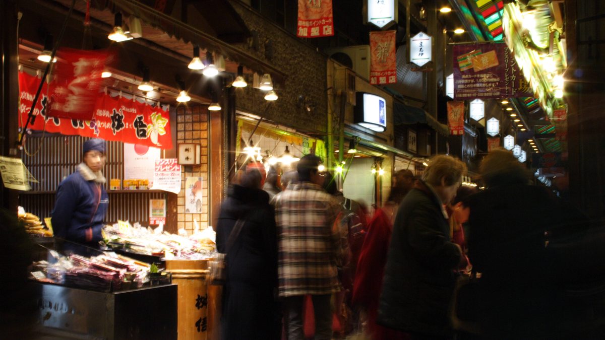 The covered arcade of Nishiki Market in Kyoto, lit by shop lanterns and busy with shoppers